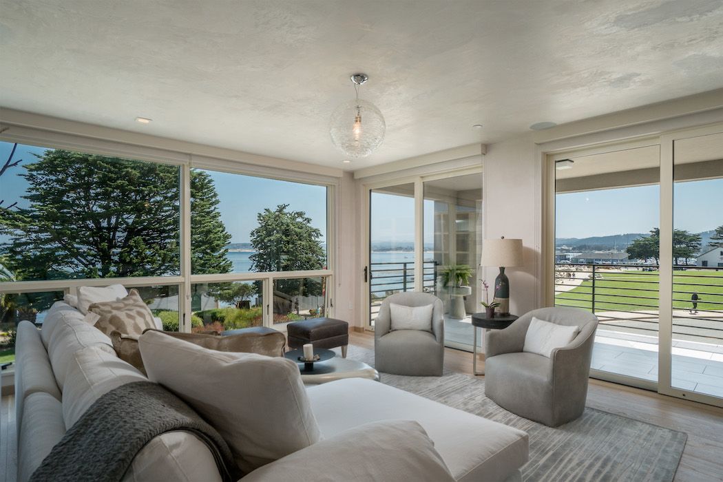 A sunlit living room with a cream sofa, two armchairs, and floor-to-ceiling windows overlooking a coastal view and lawn.