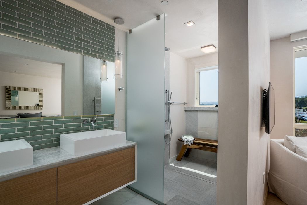 A modern bathroom features a wooden vanity with two white sinks, green subway tiles, and a frosted glass shower door.