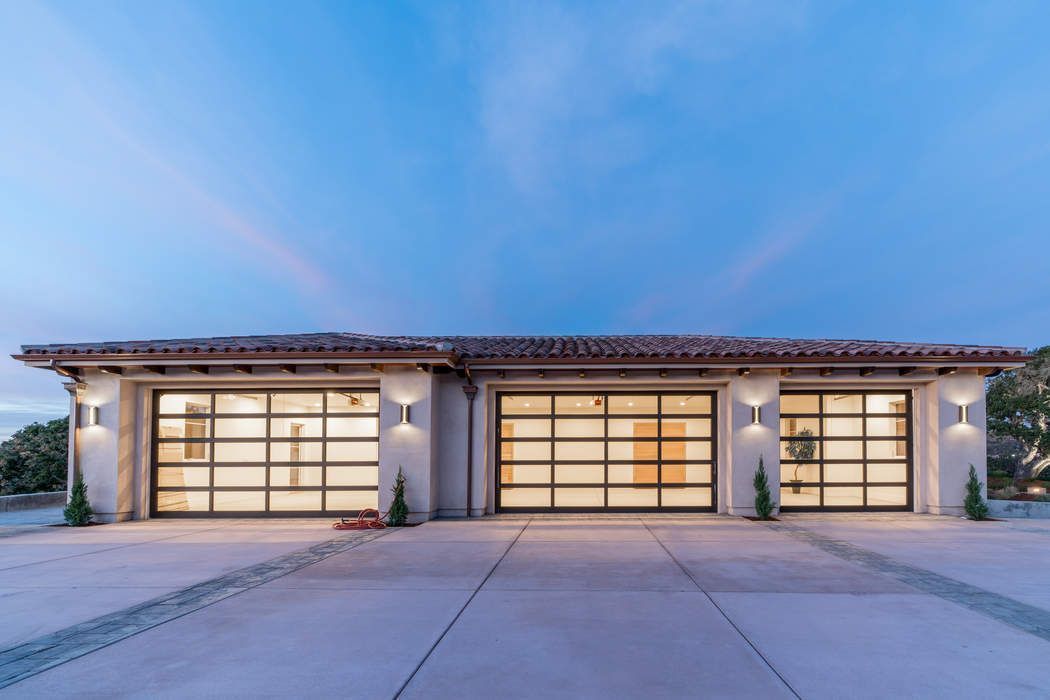 A wide shot of a three-car garage at dusk, featuring modern glass-panel doors, neutral walls, and exterior wall sconces.