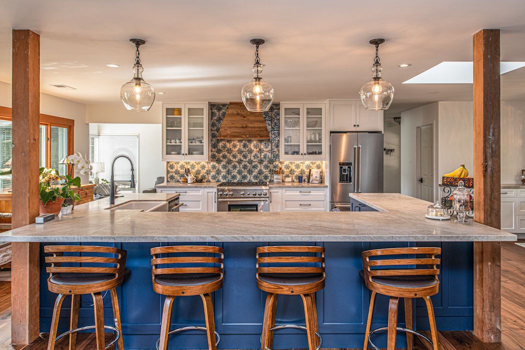 A kitchen with a large blue island, four wooden stools, white cabinets, a patterned backsplash, and three pendant lights.