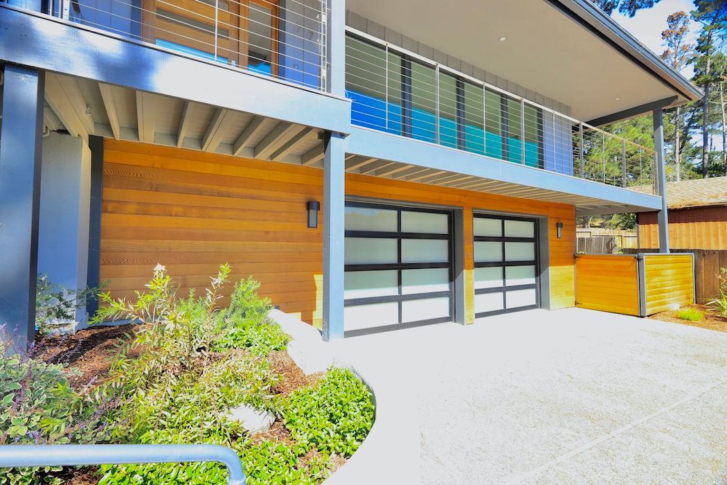 A modern home exterior with a two-car garage, wooden siding, a glass balcony, and a landscaped front garden.