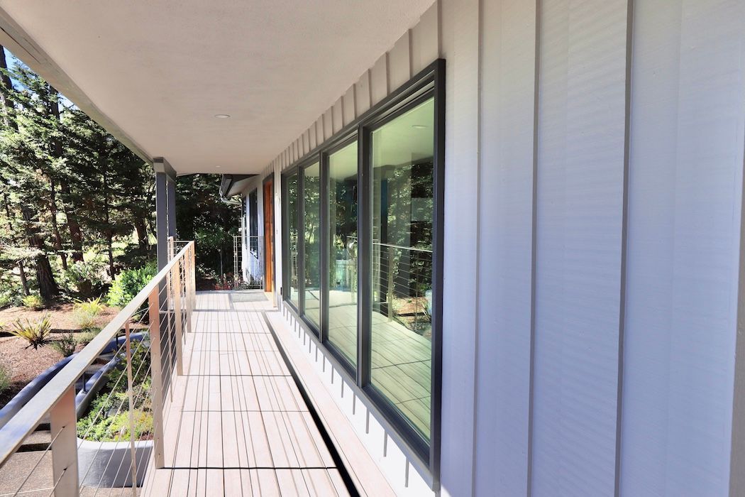 A covered deck with a railing overlooks landscaping beside a house featuring vertical light-grey siding and large windows.