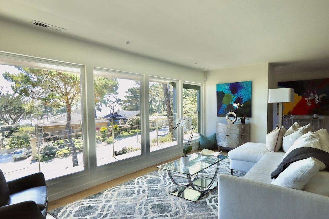 A bright, modern living room with large windows overlooking trees, a patterned rug, glass coffee table, and white sofa.