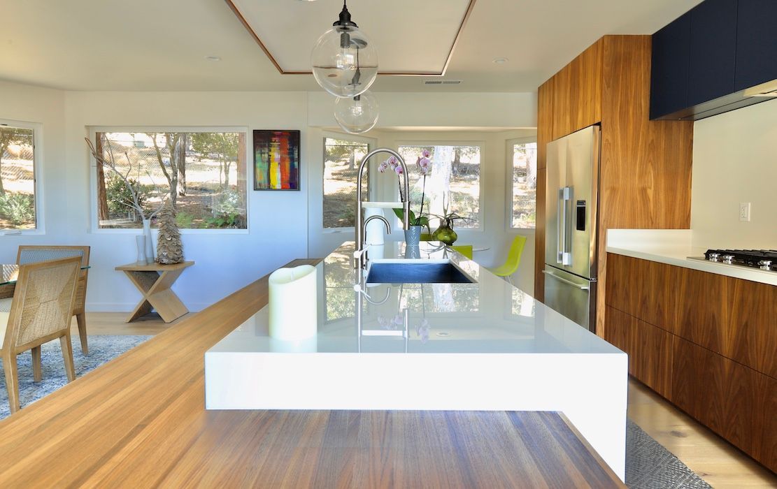 A modern kitchen island with a white countertop and wood accents, featuring a sink, stainless steel fridge, and windows.