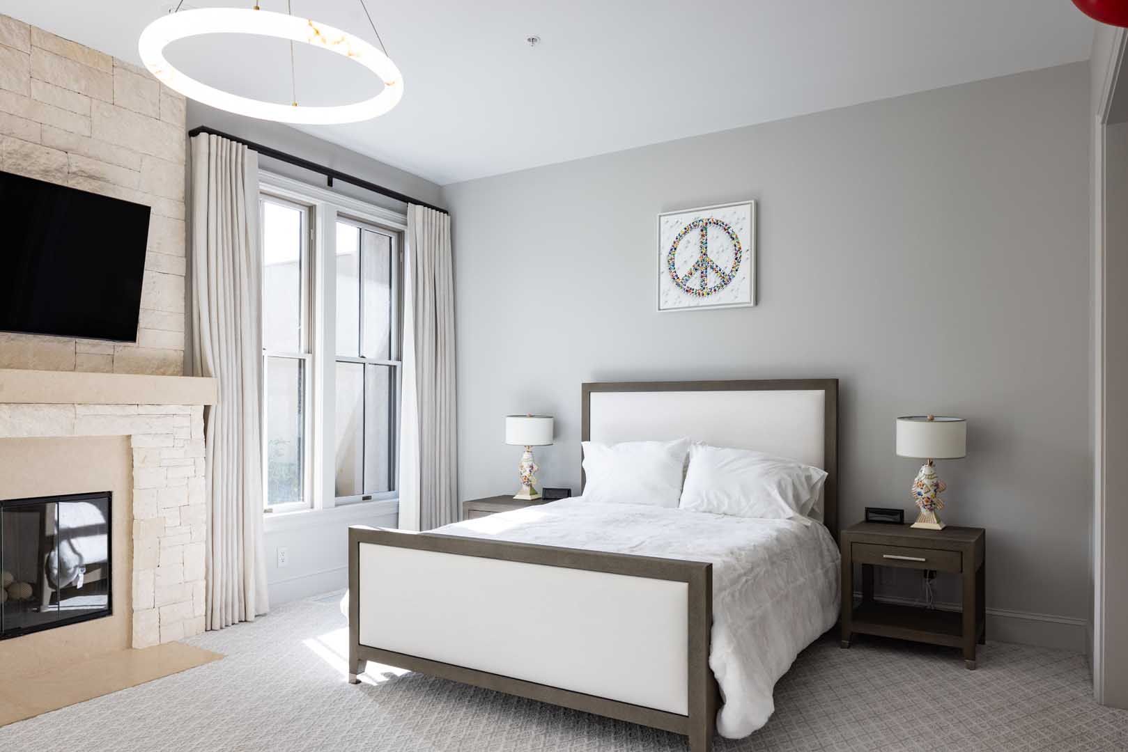 A bedroom with a bed, two nightstands, lamps, a large window with white curtains, and a stone fireplace with a TV.