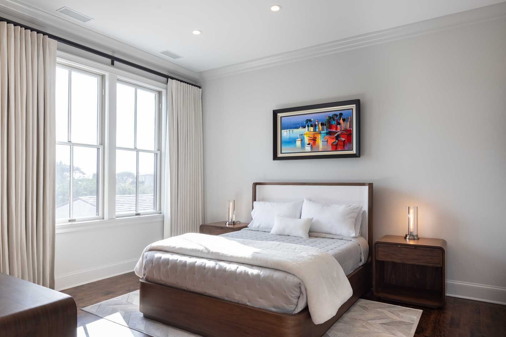 Modern bedroom with a wooden bed frame, white bedding, nightstands, floor-to-ceiling curtains, and framed wall art.