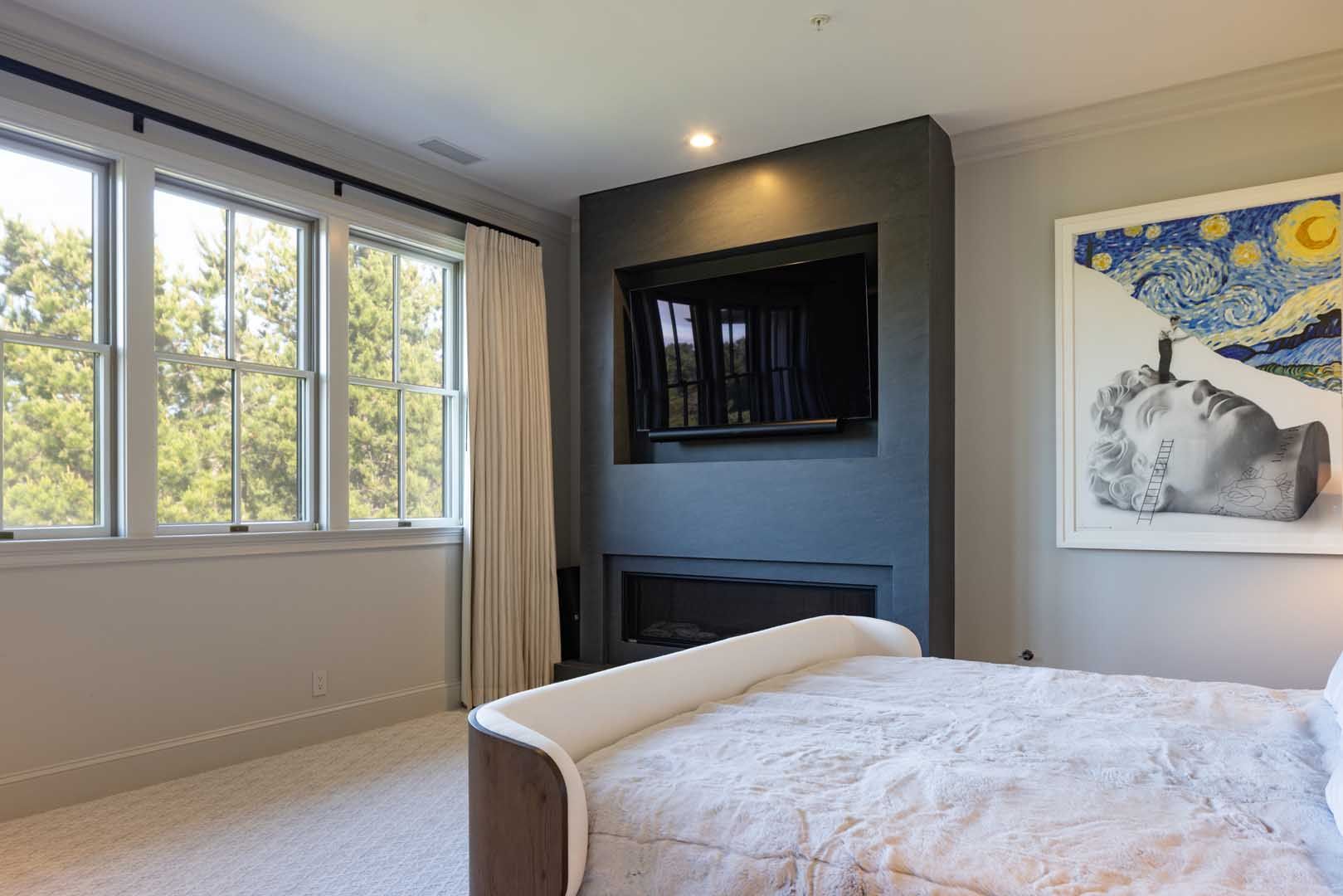 A bedroom with a bed, a dark grey fireplace wall with an integrated television, and framed art on the right.