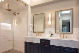 Double vanity bathroom with white marble countertops, navy blue cabinets, dual mirrors, gold fixtures, and glass shower.