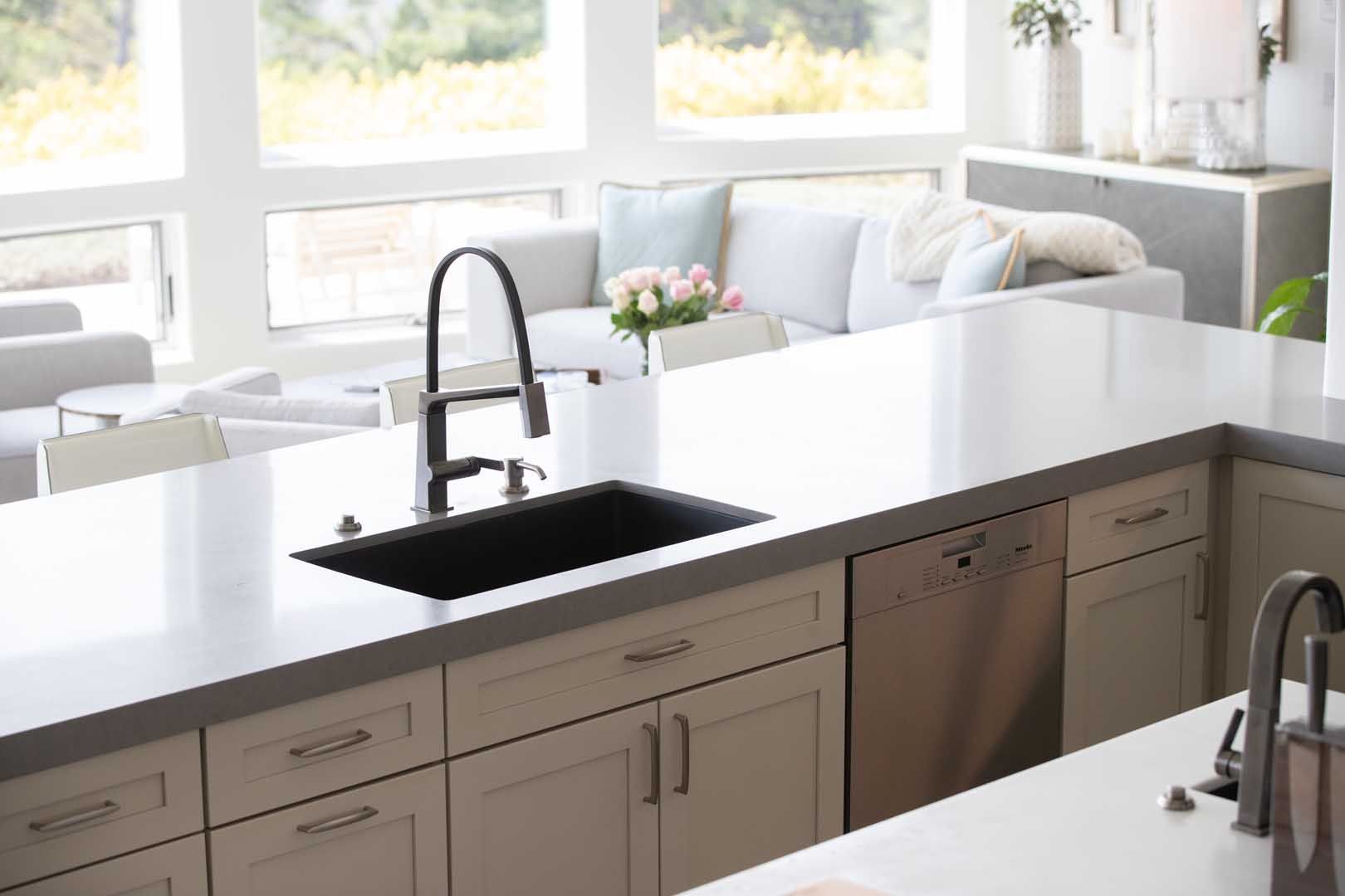A modern kitchen island with a dark sink, faucet, and dishwasher, overlooking a bright, open living room area.
