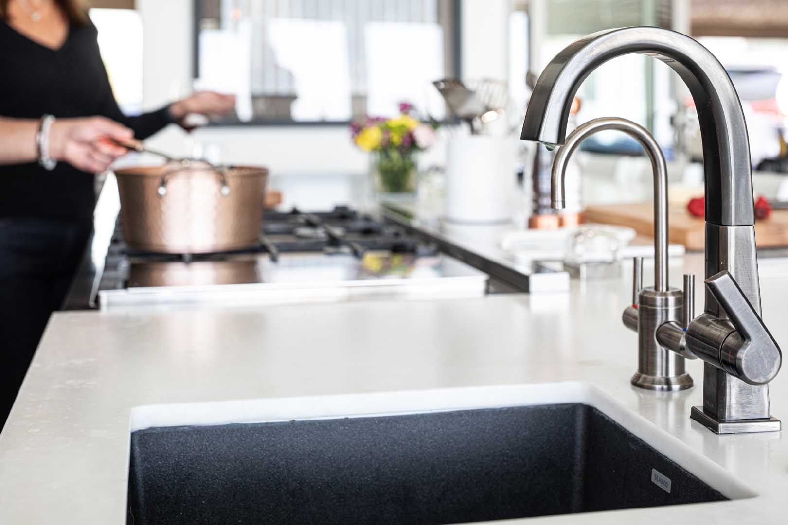 A close-up of a kitchen sink with a metal faucet and a person cooking on a stove in the blurred background.