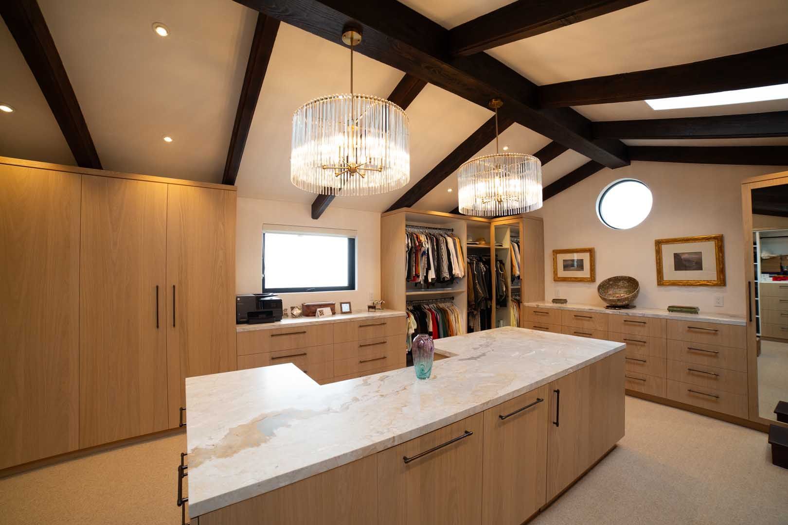 A spacious walk-in closet featuring light wood cabinetry, a marble-topped island, exposed dark beams.