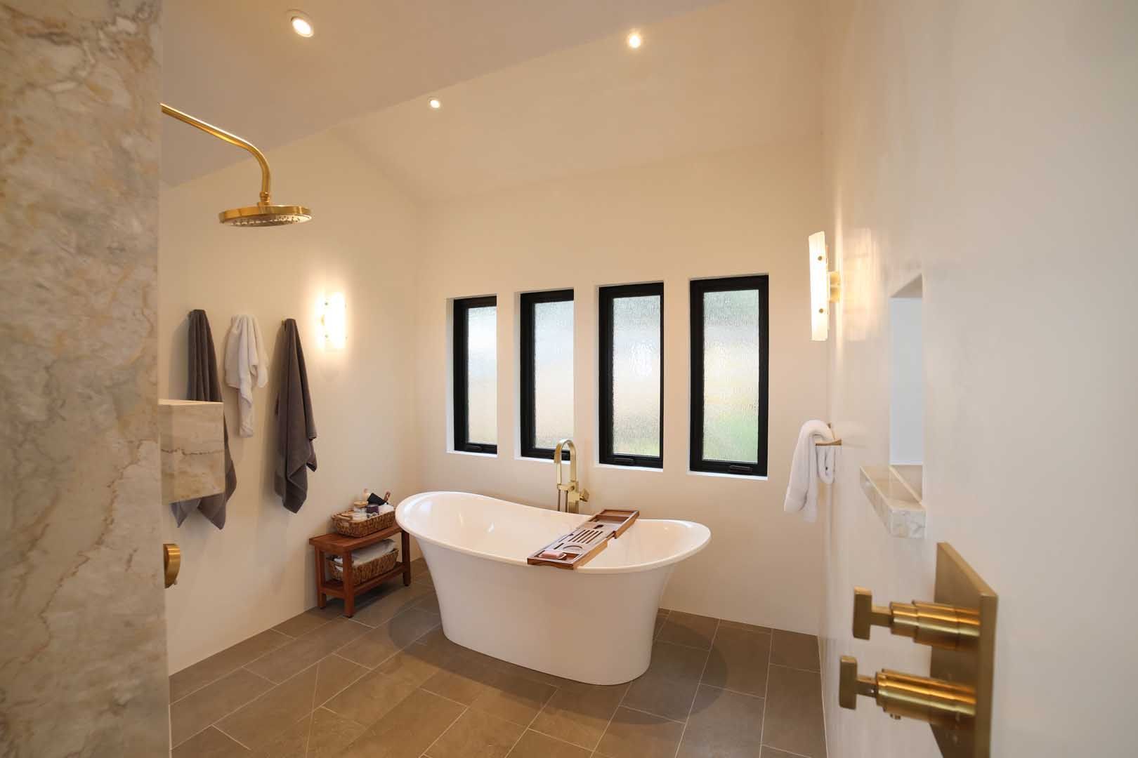A minimalist bathroom with a white freestanding tub, gold fixtures, and four vertical windows against white walls.
