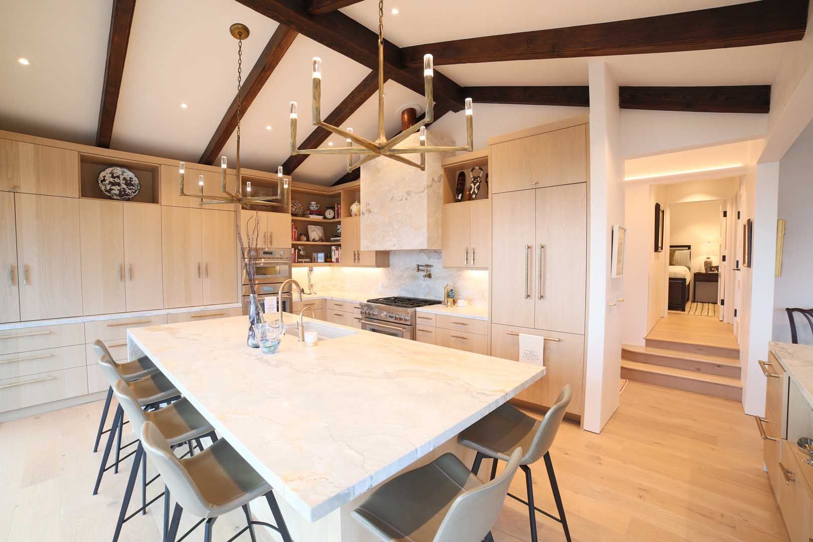 A modern kitchen with light wood cabinets, a large stone island, bar seating, and exposed dark ceiling beams.