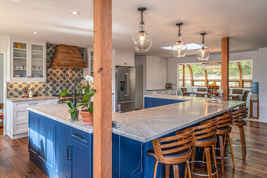 A modern, bright kitchen featuring a large blue island with a quartz countertop, wood stools, and overhead pendant lights.