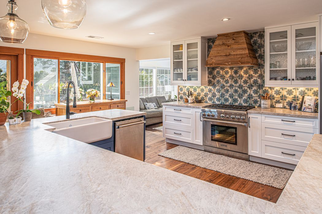 A bright, modern kitchen featuring white cabinets, a large farmhouse sink, patterned backsplash, and a wooden range hood.