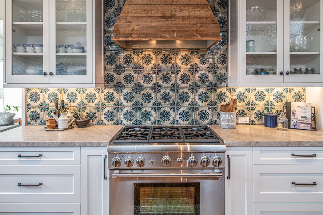 A kitchen stove with a wood range hood, blue and white patterned tile backsplash, and white cabinetry.