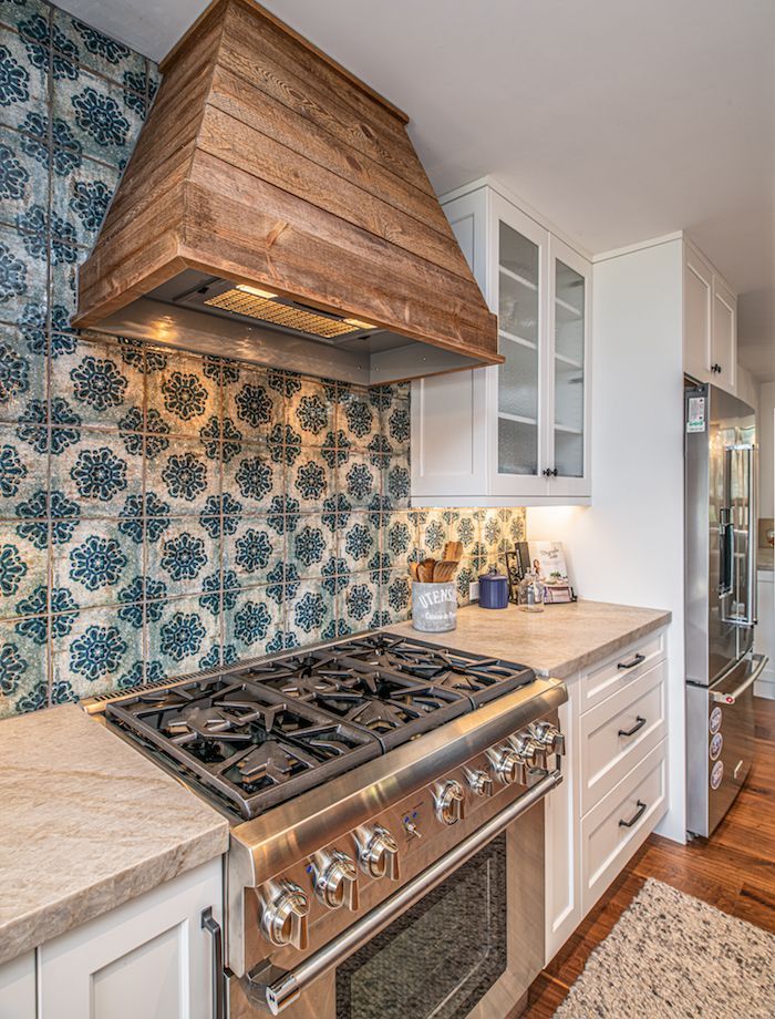 A rustic wooden vent hood above a stainless steel gas range, set against a wall with blue and white patterned tiles.