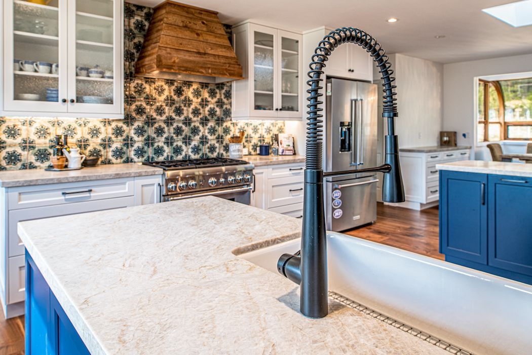 A kitchen with a large island, a stone countertop, a prominent gooseneck faucet, blue cabinetry, and patterned backsplash.