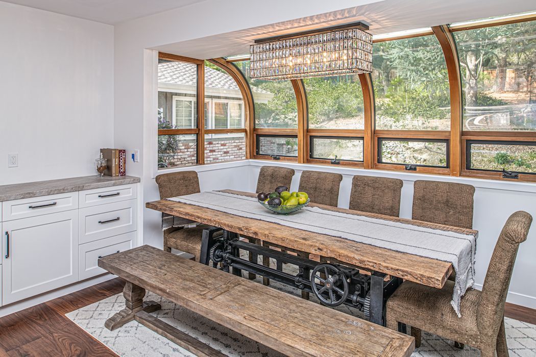 A dining nook with a long wooden table, bench, and chairs, set against a large curved window wall with a hanging light.