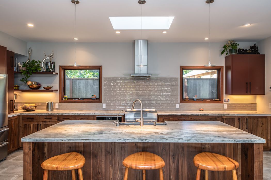 A kitchen with a large marble island, three wooden stools, dark wood cabinetry, two windows, and a stainless steel hood.