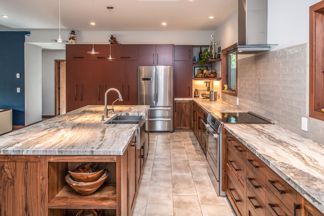 A modern kitchen with wood cabinets, a large granite island, stainless steel appliances, and beige tile flooring.