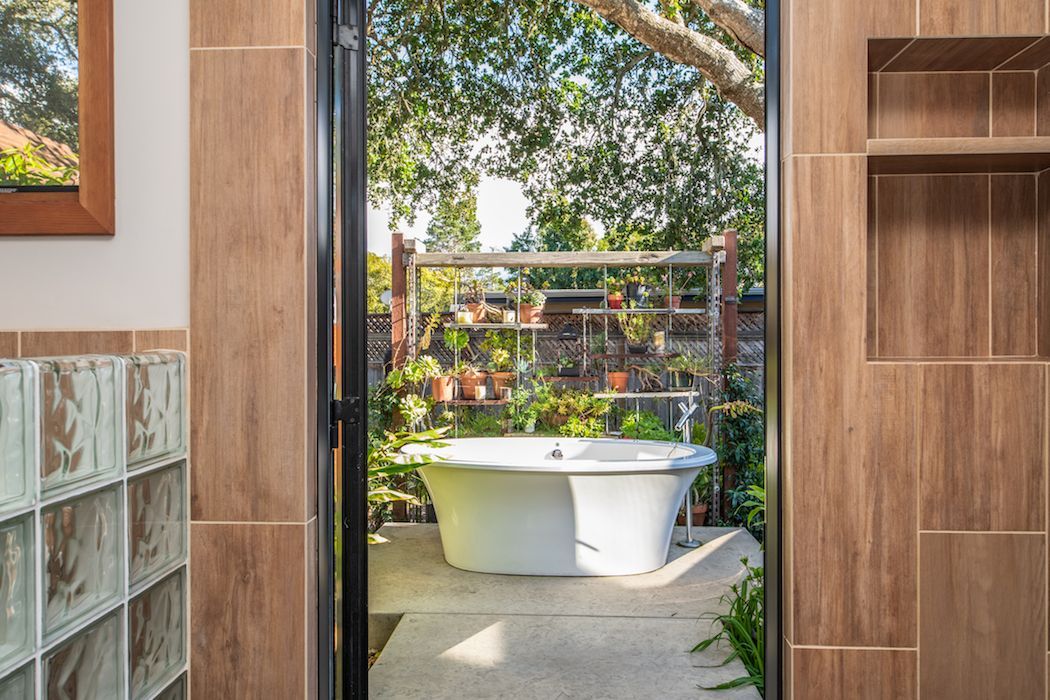 A view through a door into a lush courtyard featuring an outdoor white soaking tub surrounded by potted plants and trees.