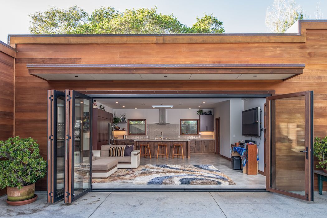 A modern wooden home features large folding glass doors opening from a patio into an interior living room and kitchen area.