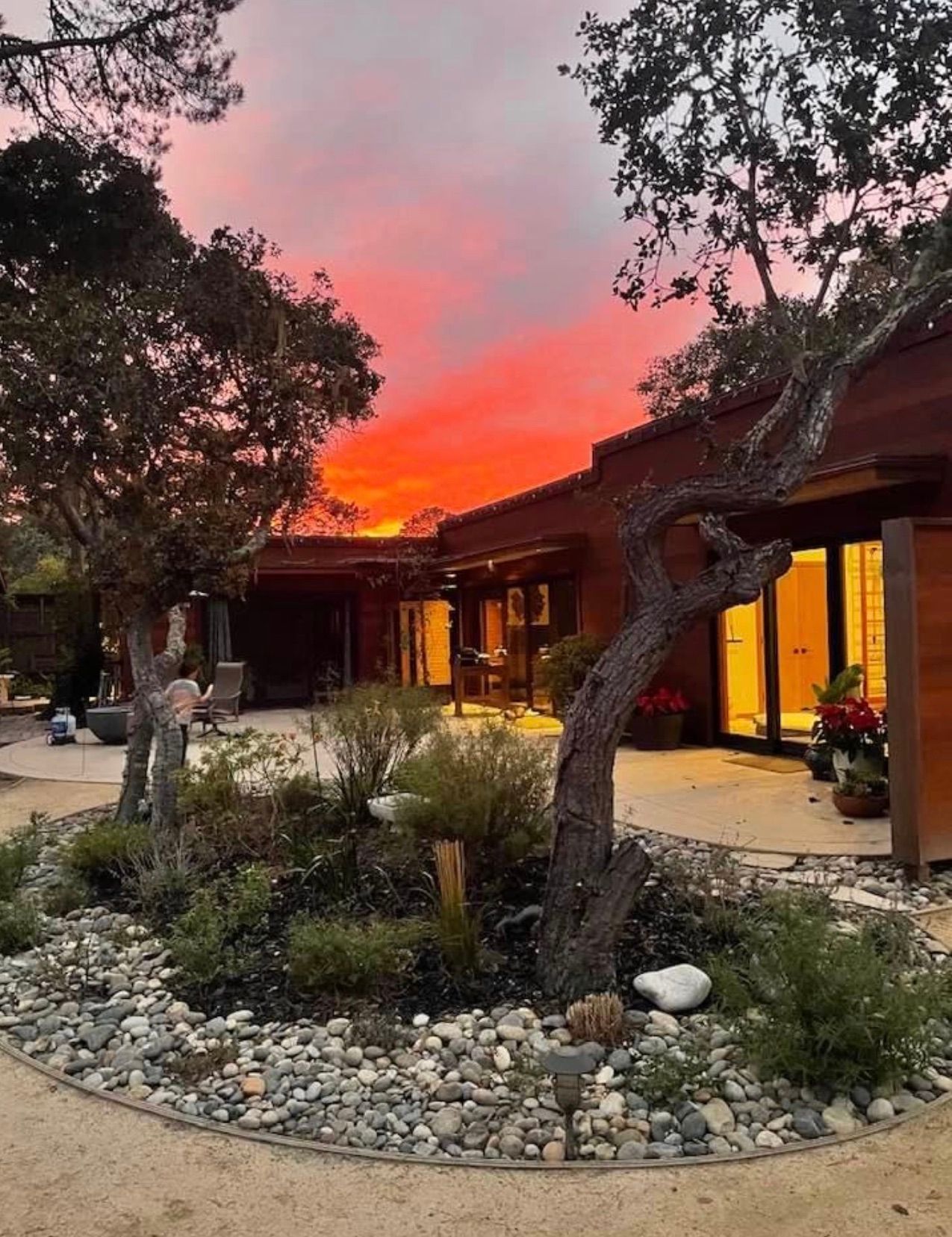 A modern home with wooden siding sits behind a circular garden with rocks and trees under a vibrant, fiery sunset sky.