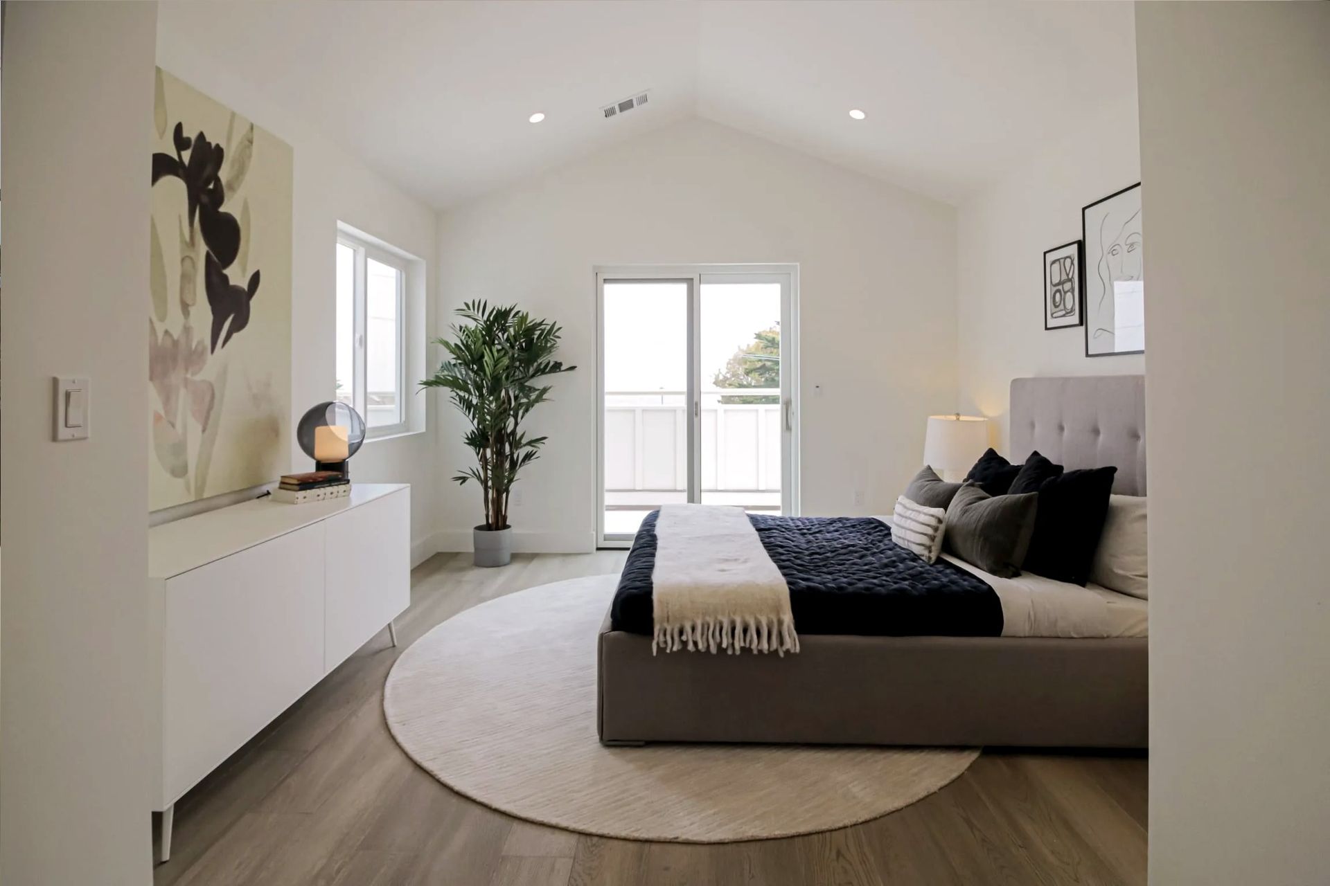 A modern, minimalist bedroom with a grey bed on a circular rug, a long white console, a potted plant, and white walls.