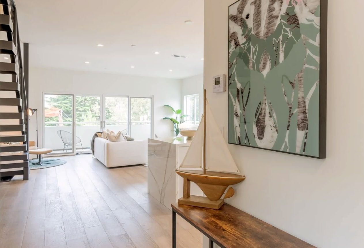 A hallway leads into a bright living room with a white sofa, past a wooden table displaying a small model sailboat.