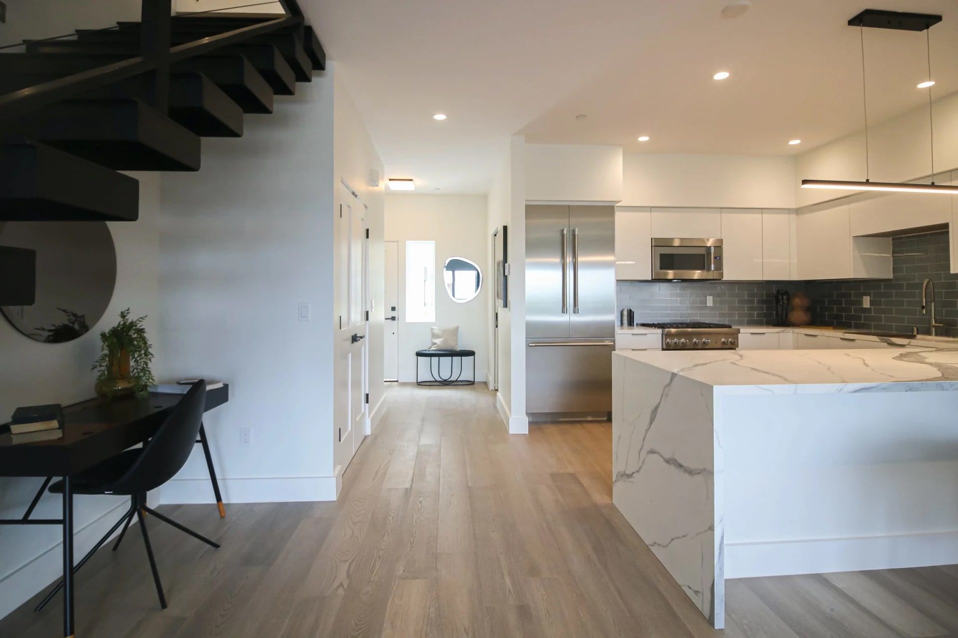 Modern open-concept interior with a white kitchen, marble island, and black staircase leading upstairs.