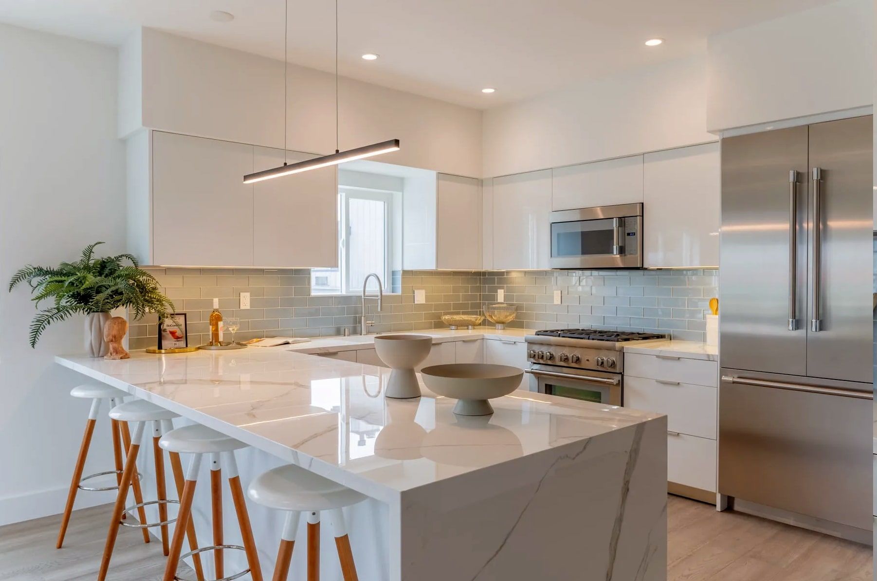 Modern kitchen featuring a white marble island with bar stools, stainless steel appliances, and gray tile backsplash.