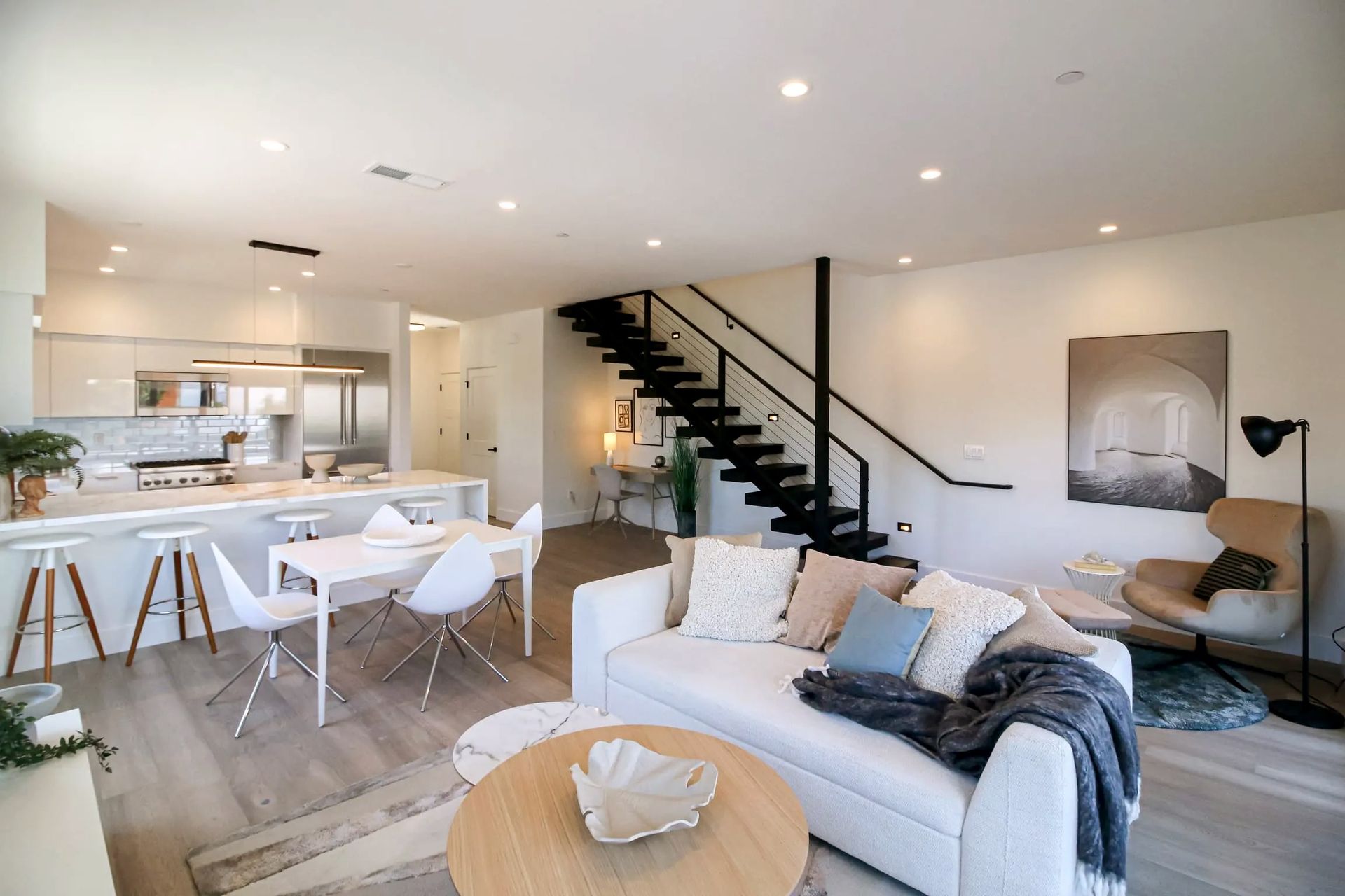 A modern open-plan living room with a white sofa, staircase, kitchen island, and dining area in a neutral-toned home.