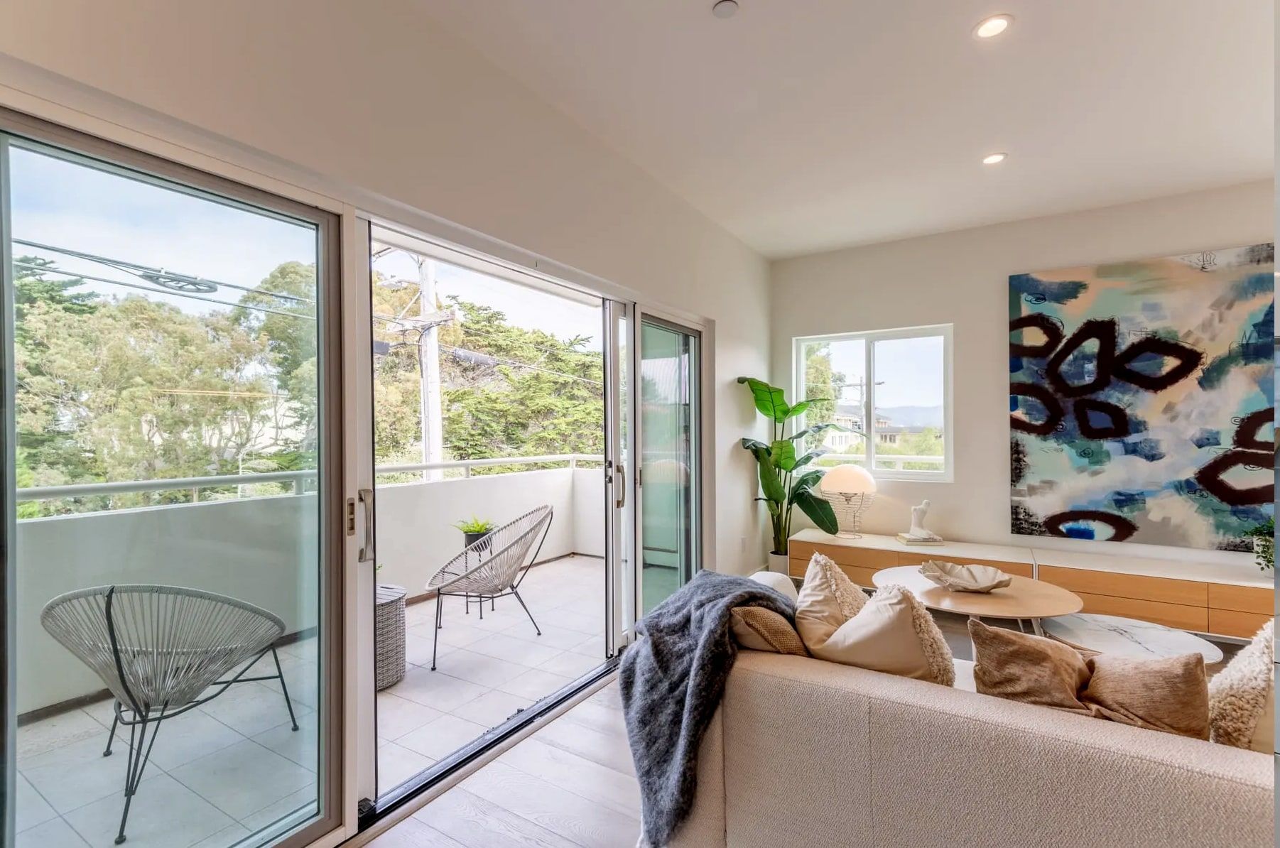 A brightly lit living room with sliding glass doors opening to a balcony featuring two metal chairs and trees outside.