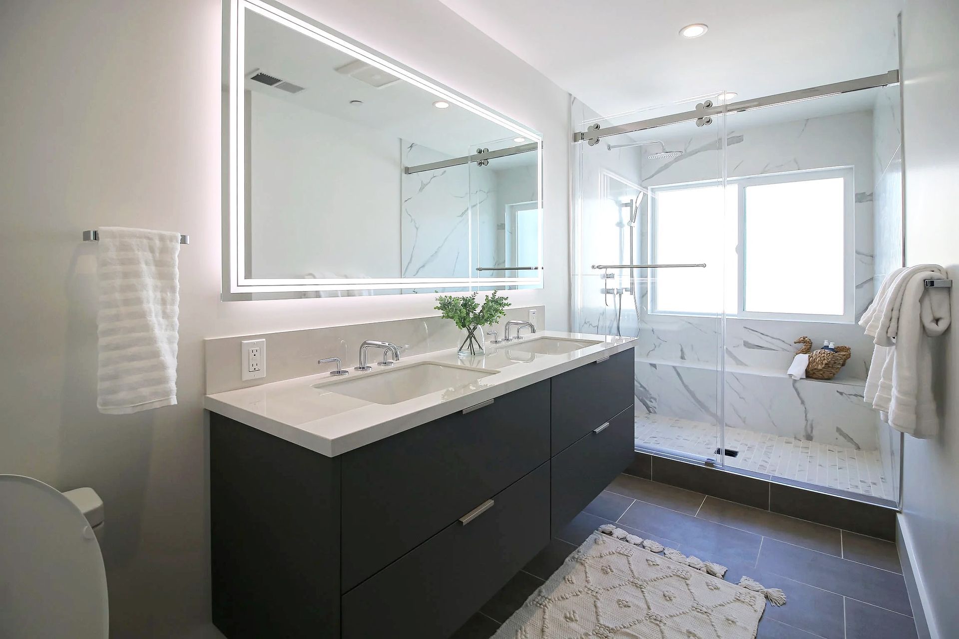 Modern bathroom with dark charcoal vanity, white marble countertops, backlit mirror, and glass-enclosed shower.