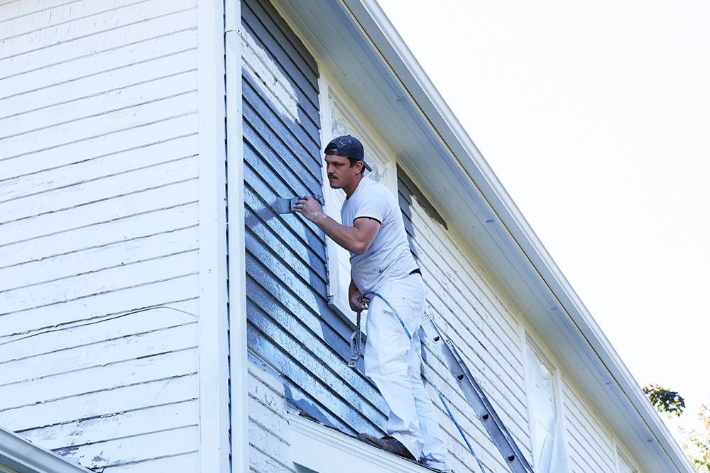 A man is standing on a ladder painting the side of a house.