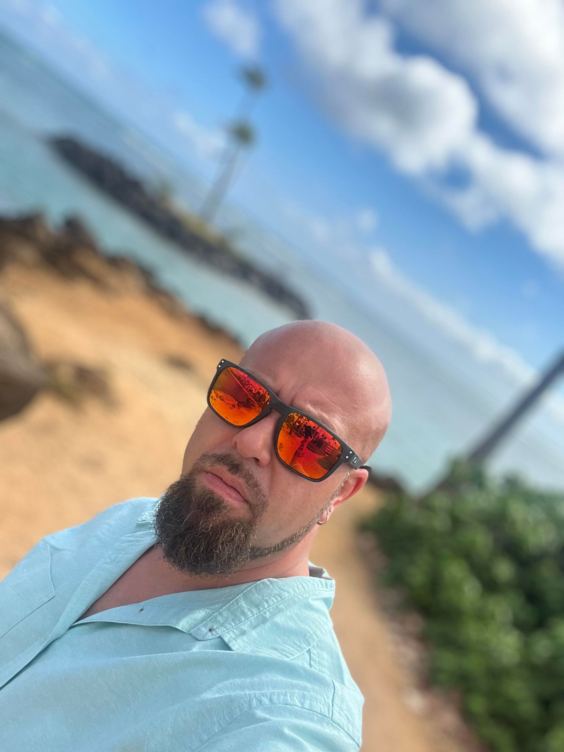 A bald man with a beard wearing sunglasses is standing on a beach.