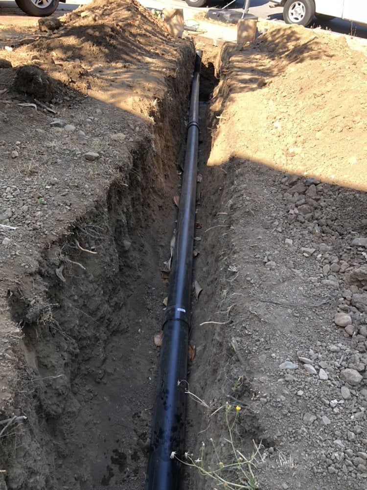 Black pipe laid in a narrow trench in the ground; dirt and gravel surround the pipe.
