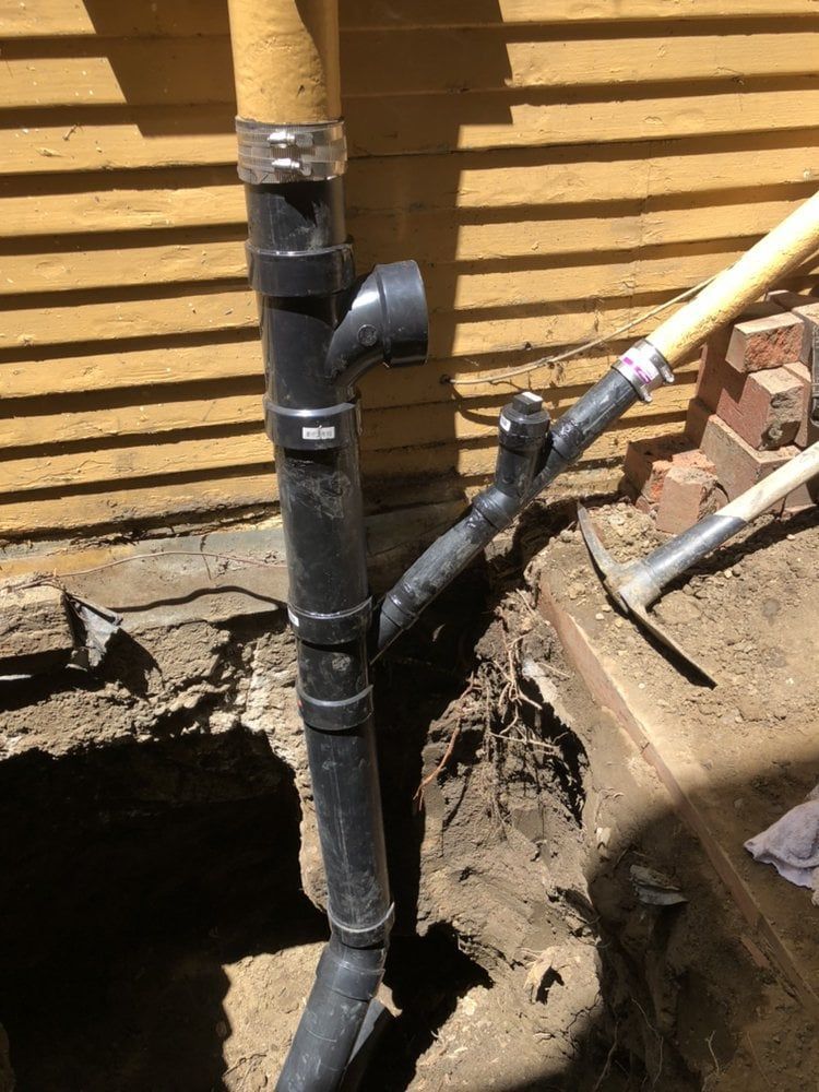 Black plumbing pipes installed in a trench against a yellow-sided wall, with shovels visible.