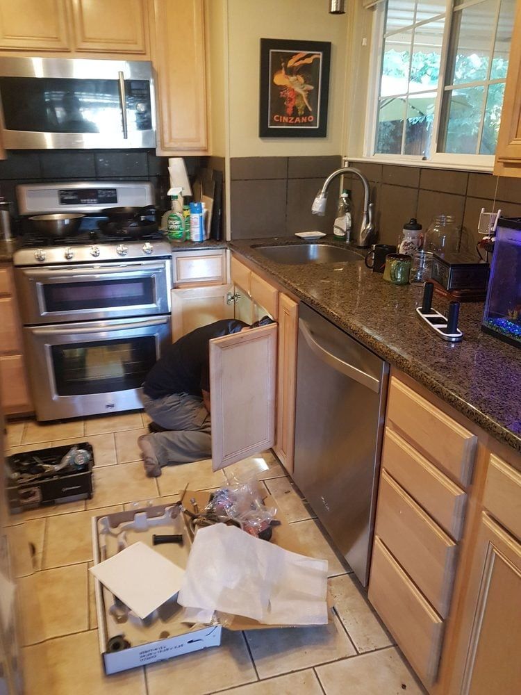 Person installing a dishwasher in a kitchen with tan cabinets, stainless steel appliances and a dark counter.