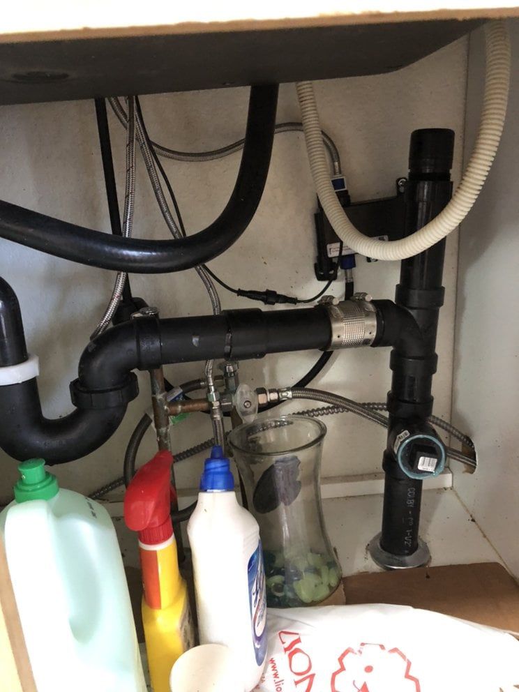 Under a kitchen sink, black plumbing pipes connect to a drain. Cleaning supplies sit on the floor.
