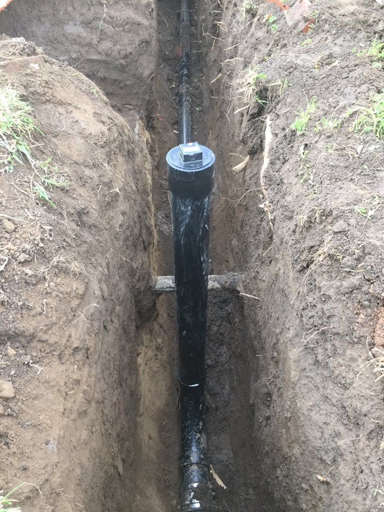 Black pipe with a large black cap installed in a narrow trench in dirt.