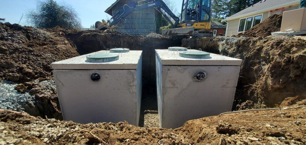 Two concrete septic tanks in a dug pit. Earth-moving equipment and a house are in the background.