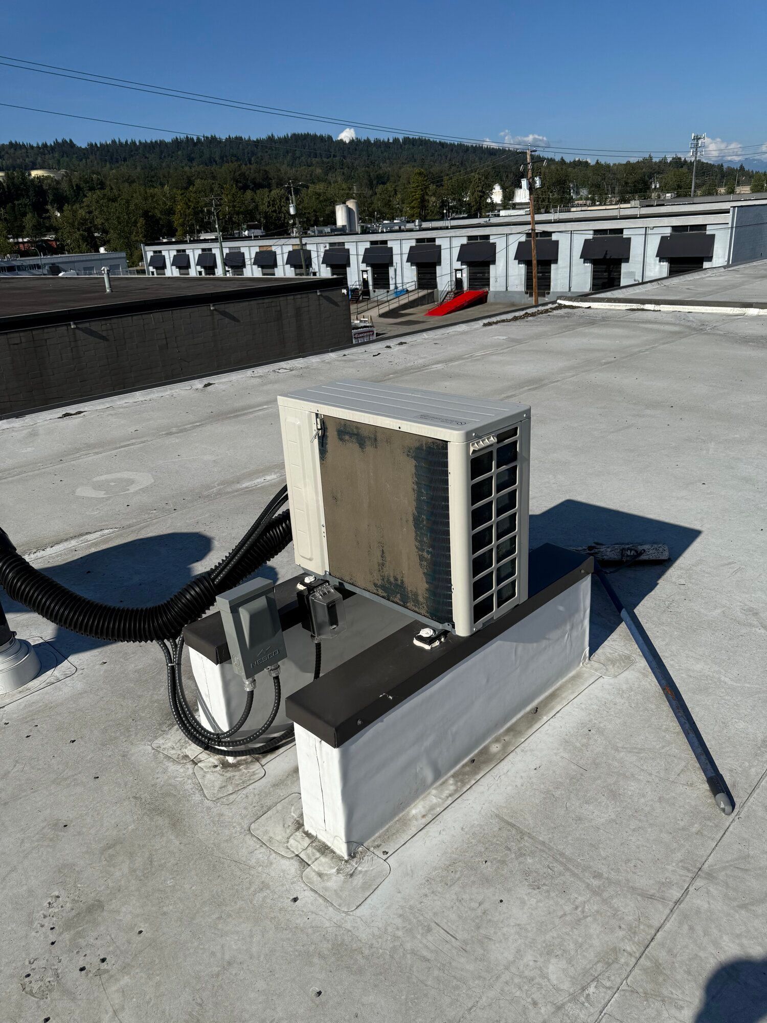 An air conditioning unit on a flat roof with a view of buildings and trees on a sunny day.