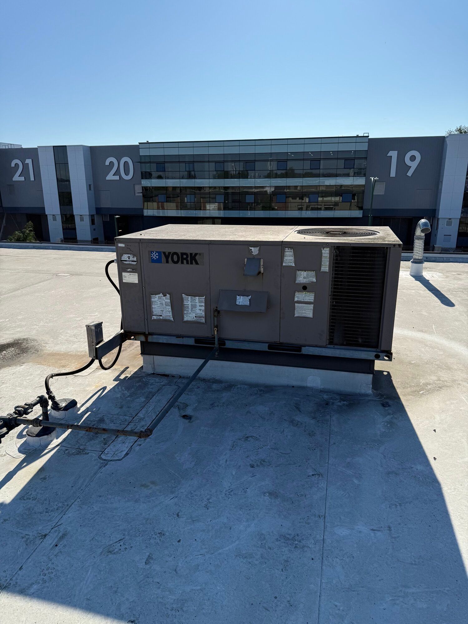 Rooftop HVAC unit; grey York machine on a flat roof, commercial building background, numbers 19-21 visible.