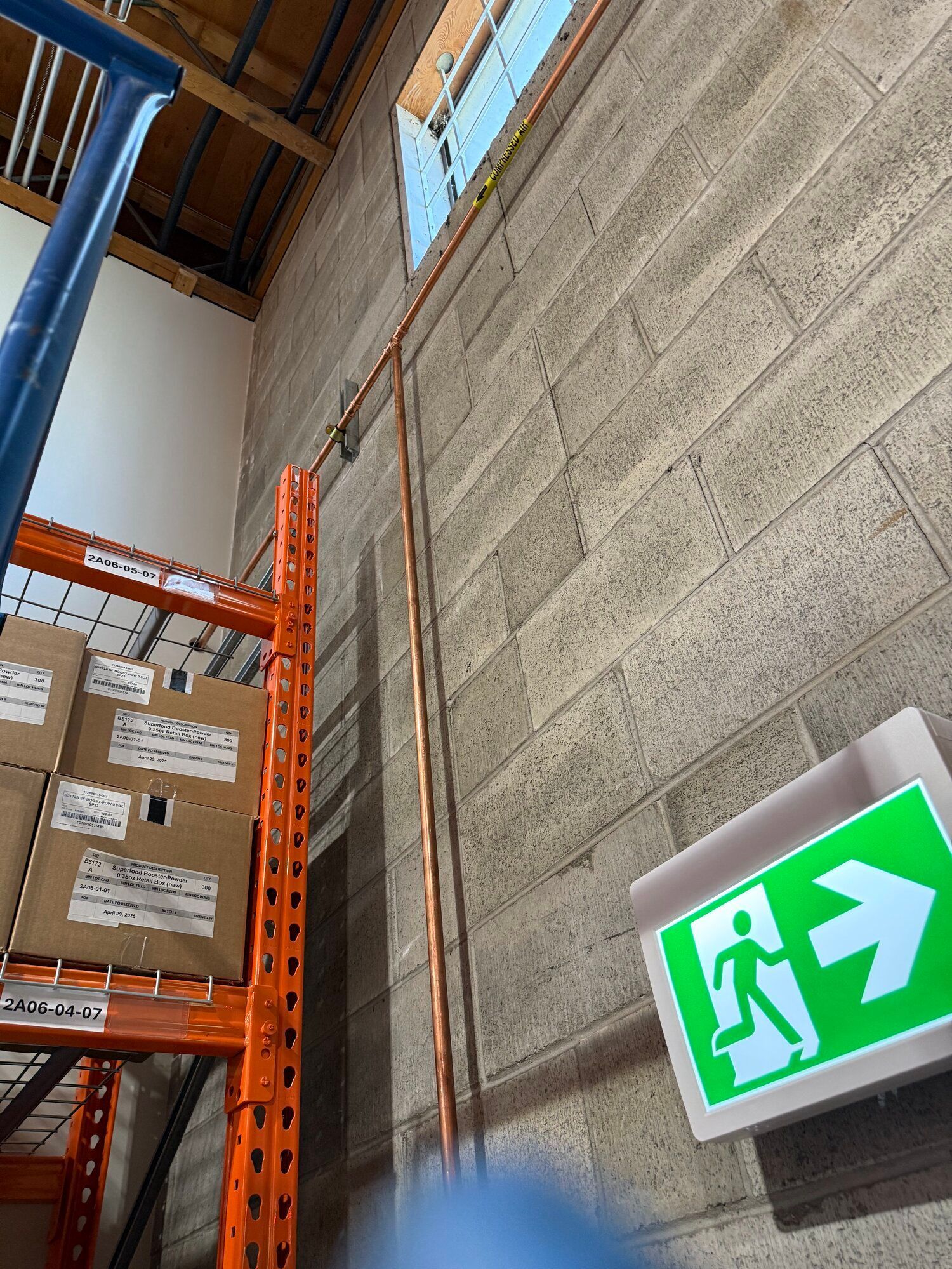 Copper pipes run vertically on a concrete block wall. An emergency exit sign is in the lower right.