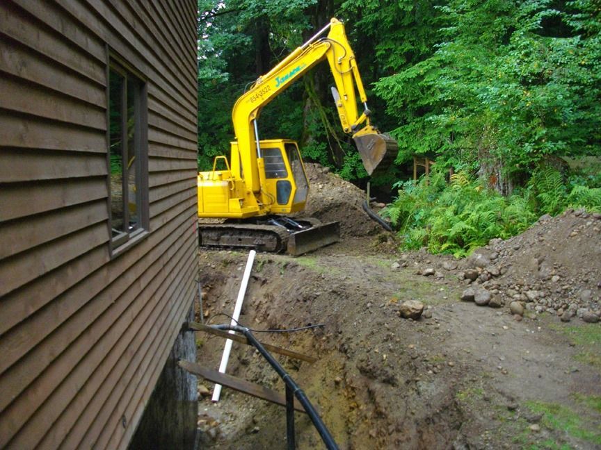 Yellow excavator digging next to a building in a wooded area.