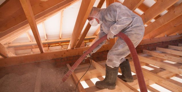 Person in protective suit spraying insulation in an attic.