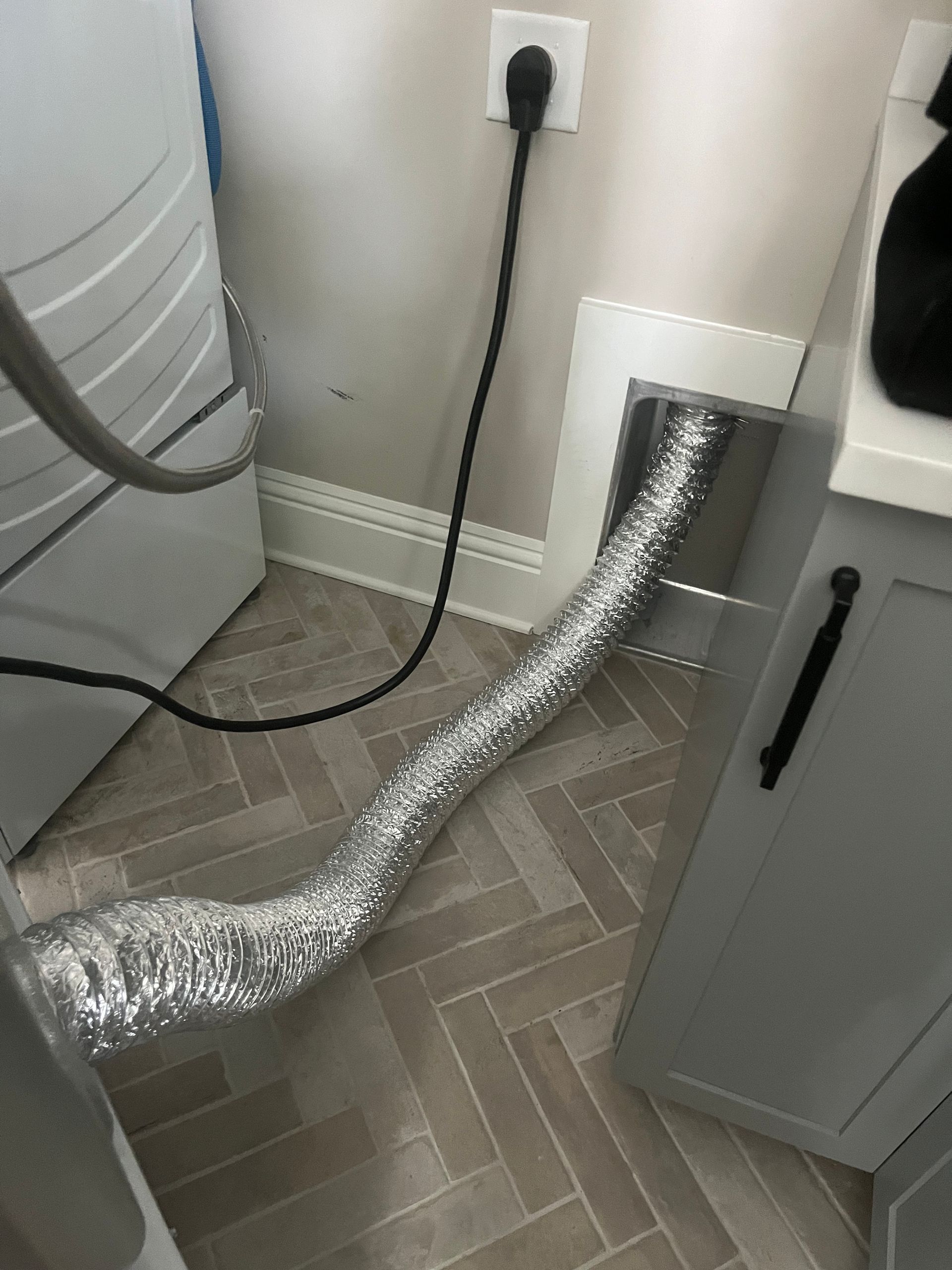 A dryer connected with a flexible vent hose to a wall vent, next to a cabinet. A power cord is plugged in.