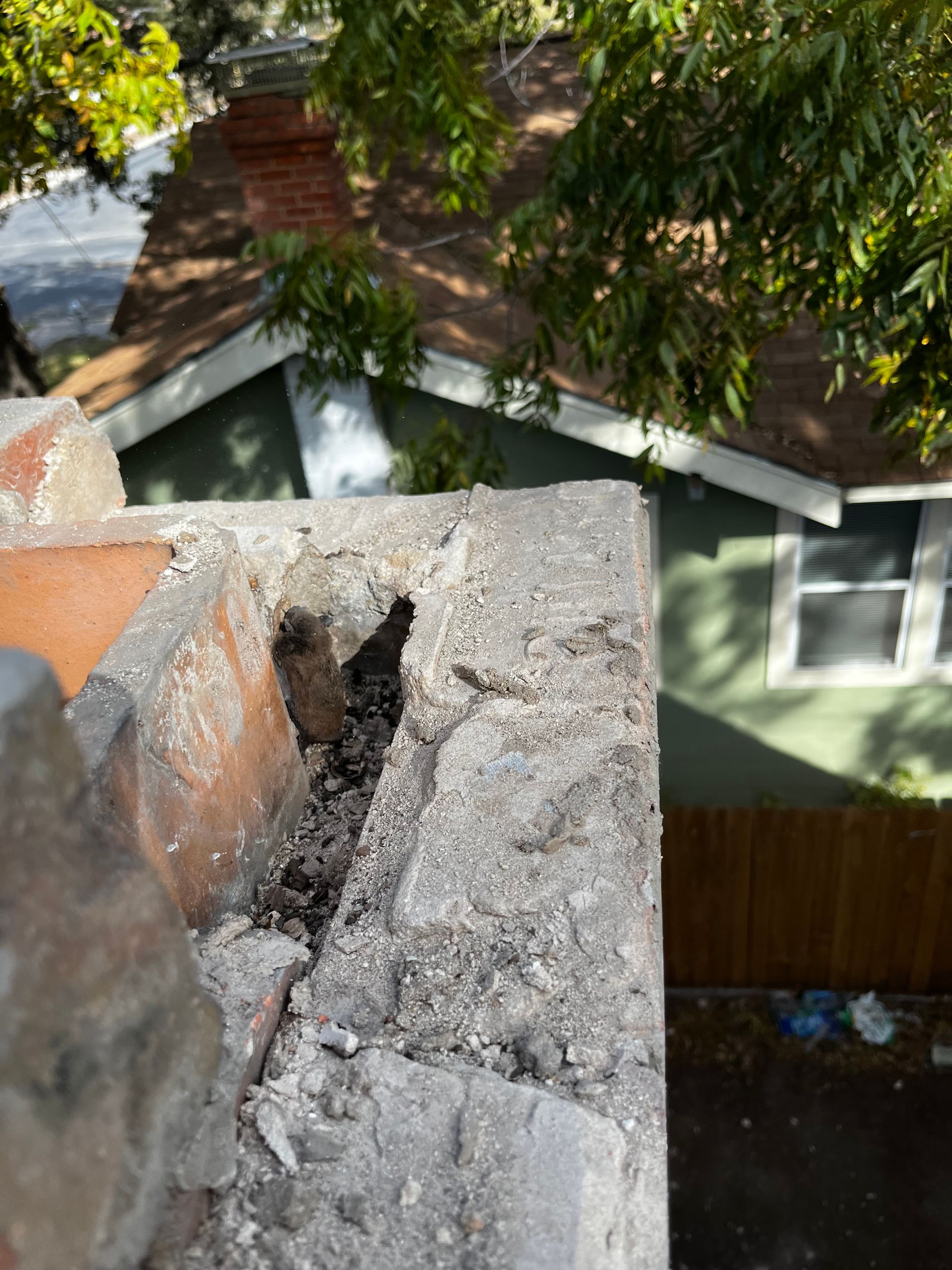 Close-up of a damaged brick chimney cap, with a deep crack exposing the interior. A house is visible in the background.